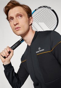 Mladý muž držící tenisovou raketu přes rameno, oblečený v černé sportovní bundě na zip Sergio Tacchini s oranžovými detaily.