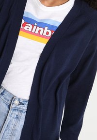 Marinblå cardigan, design med öppen front, stickat material. Bärs över en vit t-shirt med en flerfärgad "Regnbåge" grafik. Högmidjade jeanstighty synliga.