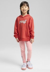 Crvena dukserica s kapuljačom i "Puma" logotipom, ružičaste tajice koje pristaju tijelu i svijetle tenisice s karikaturama. Glatka tkanina, ležeran dizajn.