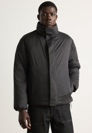 Black puffer jacket made of textured fabric, featuring a high collar, zippered front, and elastic cuffs. Worn over dark jeans.