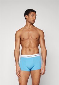 Caleçons boxers en coton bleu clair avec une taille blanche portant le logo "LACOSTE". Le design présente une forme ajustée et une texture lisse.
