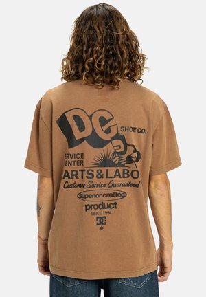 Camiseta de algodón marrón con una gran impresión gráfica negra en la parte de atrás que incluye texto y elementos de diseño. Corte holgado y mangas cortas.