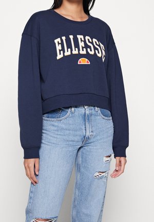 Kobieta w granatowej krótkiej bluzie z napisem i logo "ELLESSE", w zestawieniu z jasnoniebieskimi, podartymi, wysokim stanem jeansami.