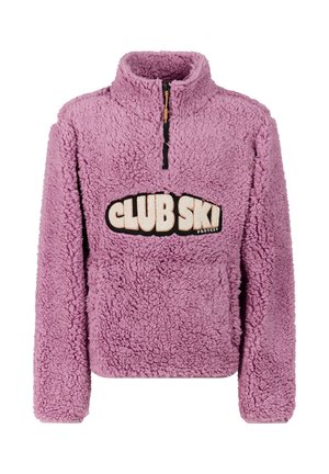Junge Frau trägt einen lila Fleecepullover mit dem Text "CLUB SKI", Leopardenmuster-Hose, rosa Mütze und schwarze Schuhe, steht mit den Händen in den Taschen.
