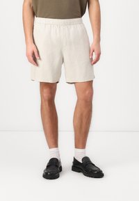 Beige linshorts med elastisk midje, med sidelommer og løs passform, kombinert med svarte slip-on sko og hvite sokker.