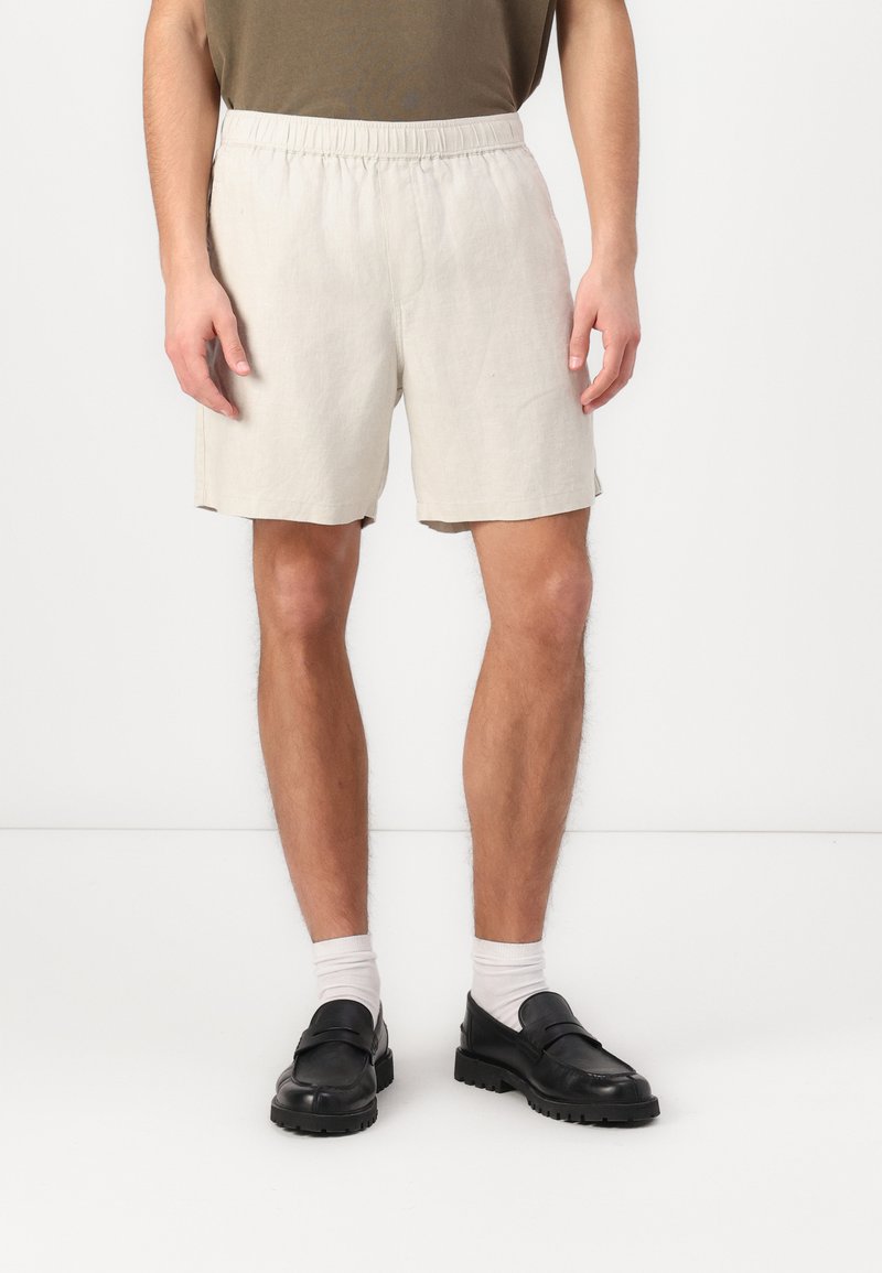 Shorts en lin beige avec une taille élastique, dotés de poches latérales et d'une coupe décontractée, associés à des chaussures noires à enfiler et des chaussettes blanches.