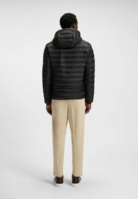 Giacca a vento nera con cappuccio, caratterizzata da una texture trapuntata e polsini elasticizzati, abbinata a pantaloni beige a gamba affusolata e scarpe nere.