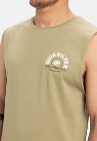 Kreklu bez piedurknēm, olīvu zaļā krāsā, izgatavots no mīkstas kokvilnas. Tam ir balta grafiskā logo ar tekstu "QUIKSILVER THE ORIGINAL BOARDRIDING" priekšpusē.