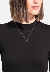 Collana con pendente a forma di cuore in oro, indossata su una maglia a collo alto nera, che mette in risalto il design minimalista e la texture liscia.