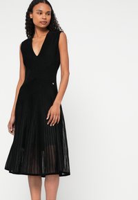 Μαύρο αμάνικο φόρεμα με V-ντεκολτέ, διαθέτοντας κάθετη ριγέ υφή και φουσκωτό φούστα. Λεπτομέρεια με κουμπί στη μέση.