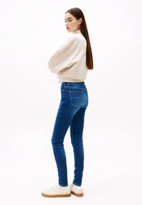 Moteris stovinti sukryžiuotomis rankomis, vilkinti smėlio spalvos džemperį, mėlynas aptinpias džinsus, baltus sportinius batelius, ilgus tiesius rudus plaukus, prieš baltą foną.