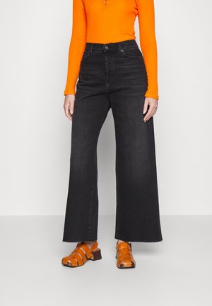 Personne portant un jean noir taille haute à jambes larges, un haut orange à manches longues et des sandales orange à brides avec talons blocs, debout sur un fond uni.