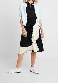 Světlomodrý svetr přes černý top, plisovaná sukně v černé a krémové barvě a bílé tenisky. Texturovaná látka, vrstvený vzor, volný střih.