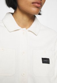 Vit cordflorskjorta med krage, utrustad med två knäppbara bröstfickor och en svart etikett med "OBEY" på vänster ficka.