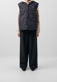 Chaleco negro acolchado con cuello y botones a presión, llevado sobre una sudadera beige. Pantalones sueltos negros a rayas y zapatos negros de punta.