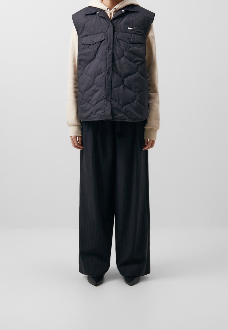 Chaleco negro acolchado con cuello y botones a presión, llevado sobre una sudadera beige. Pantalones sueltos negros a rayas y zapatos negros de punta.