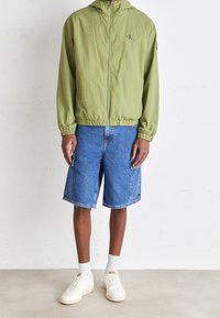 Person som bär en ljusgrön zip-upp-jacka, blå denimshorts med sidofickor, vita strumpor och off-white sneakers, stående på en vanlig golv.