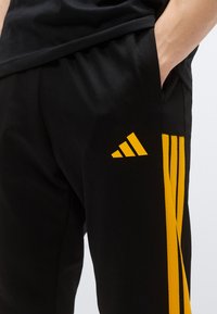 Μαύρο αθλητικό παντελόνι με υφασμένο μαύρο ρίγα και κίτρινο λογότυπο Adidas στο πλάι. Κομμένο με κομψό, στενό σχεδιασμό και ευρύχωρη τσέπη.