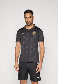 Camisola de futebol preta com um padrão subtil do brasão de armas de Portugal, logótipo dourado e gola às riscas vermelhas e verdes. Usada com calções pretos.