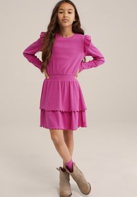 Rosa långärmad klänning med volangdetaljer på axlarna och en lager-på-lager nederkant. Texturerat tyg, elastiskt midjeband, matchad med beige ankelstövlar.