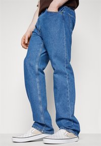 Blue denim jeans with a straight-leg fit, featuring side pockets and faint whiskering details. Paired with white checkered slip-on shoes.