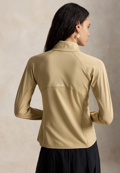 Γυναίκα με μακριά σκούρα μαλλιά, φοράει μπεζ μπουφάν με φερμουάρ και λογότυπο RLX στην πλάτη και μαύρη φούστα, εμφανίζεται από πίσω με ουδέτερο φόντο.