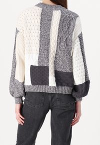 Pull en tricot gris et crème avec un design en patchwork, texture chunky et poignets côtelés, offrant une coupe décontractée.