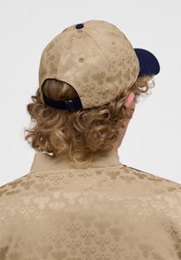 Beige Kappe mit einem dunkelblauen Visier, ausgestattet mit einem durchgehenden geprägten Muster aus stilisierten Motiven. Mit Klettverschluss auf der Rückseite.
