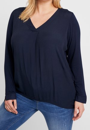 Marineblaues Langarmshirt mit V-Ausschnitt und Frontfalte; weiche Textur, lockere Passform, detaillierte Manschetten, kombiniert mit hellblauen Jeans.