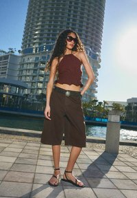Haut halter court marron, pantalon large marron avec une ceinture ornée d'un accent turquoise, et sandales noires à brides; cadre urbain au bord de l'eau.