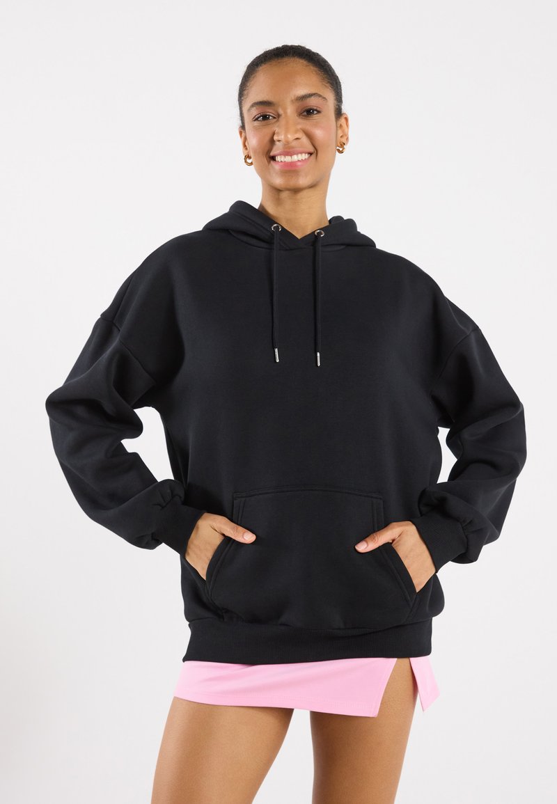 Hoodie oversize nero con cappuccio regolabile, tasca a marsupio frontale e polsini a costine. Indossato sopra una gonna rosa. Tessuto dalla superficie liscia.