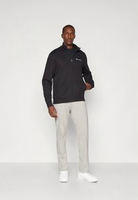 Zwarte fleecejas met ritssluiting en opstaande kraag, gedragen over een grijs T-shirt en lichtgrijze joggingbroek. Inclusief witte sneakers.