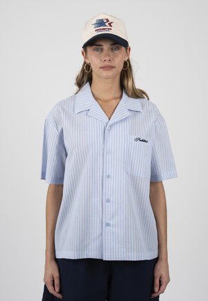 Kobieta ubrana w jasnoniebieską, krótko rękawową, w paski koszulę zapinaną na guziki i beżową czapkę baseballową z logo, stojąca na jednolitym tle.