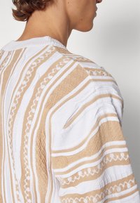 Stickad tröja i vitt och beige med randiga mönster. Har texturerade tyger och en ribbad krage. Öppen stickad design på baksidan.
