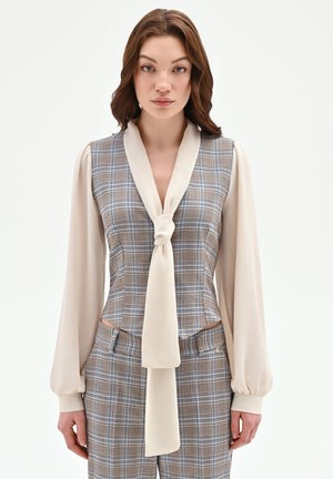 Femme portant un chemisier beige à manches longues avec un foulard noué autour du cou sous un gilet à carreaux marron et bleu, assorti à un pantalon à carreaux, debout immobile.