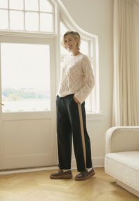 Jasnoróżowy sweter dzianinowy z teksturowanym wzorem w diamenty, w połączeniu z czarnymi spodniami z beżowym paskiem po boku i brązowymi butami.