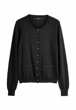 Frau mit langen braunen Haaren trägt eine schwarze geknöpfte Strickjacke, weiße Hosen und goldene Ohrringe, steht vor einem schlichten Hintergrund.