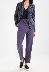 Námorní blazer s potiskem a hladkou texturou, spárovaný s volnými fialovými kalhotami s opaskem na zavázání a černými klouzavými botami.