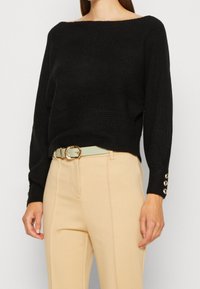 Pull noir en maille avec un col bateau, des manches longues et une texture côtelée. Associé à un pantalon beige et une ceinture de couleur claire avec des accents dorés.