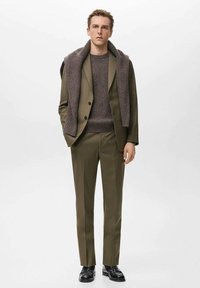 Maslinasto zeleni odijelo krojeno po mjeri s teksturiranim smeđim džemperom i pletenim šalom preko ramena. Crne kožne cipele upotpunjuju odjevnu kombinaciju.