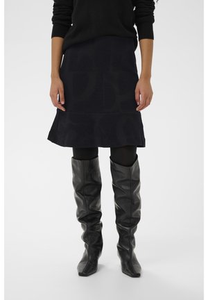 Vrouw draagt een zwarte, getextureerde rok, zwarte panty's, een zwarte trui en losse zwarte leren kniehoge laarzen, staand voor een effen achtergrond.