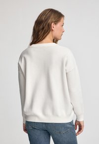 Weißer Baumwollpullover mit lässigem Schnitt, geripptem Halsauschnitt und Bündchen. Kombiniert mit blauen Jeans aus Denim hat der Stoff eine weiche und glatte Textur.