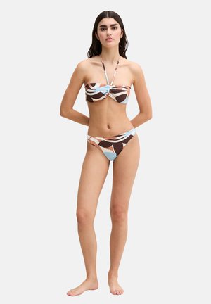Kobieta stojąca boso, ubrana w bikini bez ramiączek z abstrakcyjnym, wielokolorowym wzorem i wiązaniem na szyi, na jednolitym białym tle.