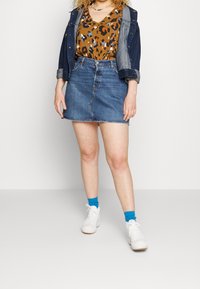 Jupe en denim avec un ourlet légèrement effiloché, associée à un haut à imprimé léopard marron et une veste en denim. Des baskets blanches et des chaussettes bleues complètent le look.