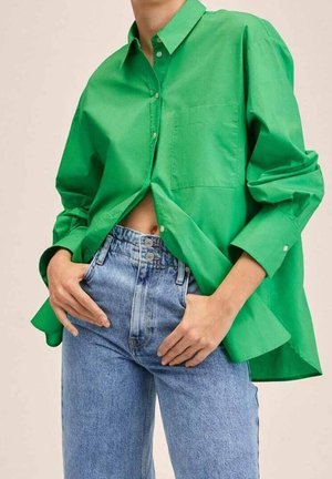 Person iført en oversized, knaldgrøn skjorte og lyseblå højtaljede jeans med hænderne hvilende nær lommerne.