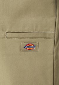 Un pantalon de travail beige avec une poche arrière dotée d'une étiquette. L'étiquette présente un logo circulaire multicolore avec "Dickies" écrit en texte bleu. Tissu texturé.