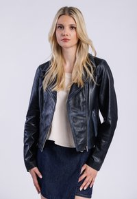 Blondynka w czarnej skórzanej kurtce i jasnej bluzce, stojąca na jednolitym białym tle, z pewnym siebie wyrazem twarzy.