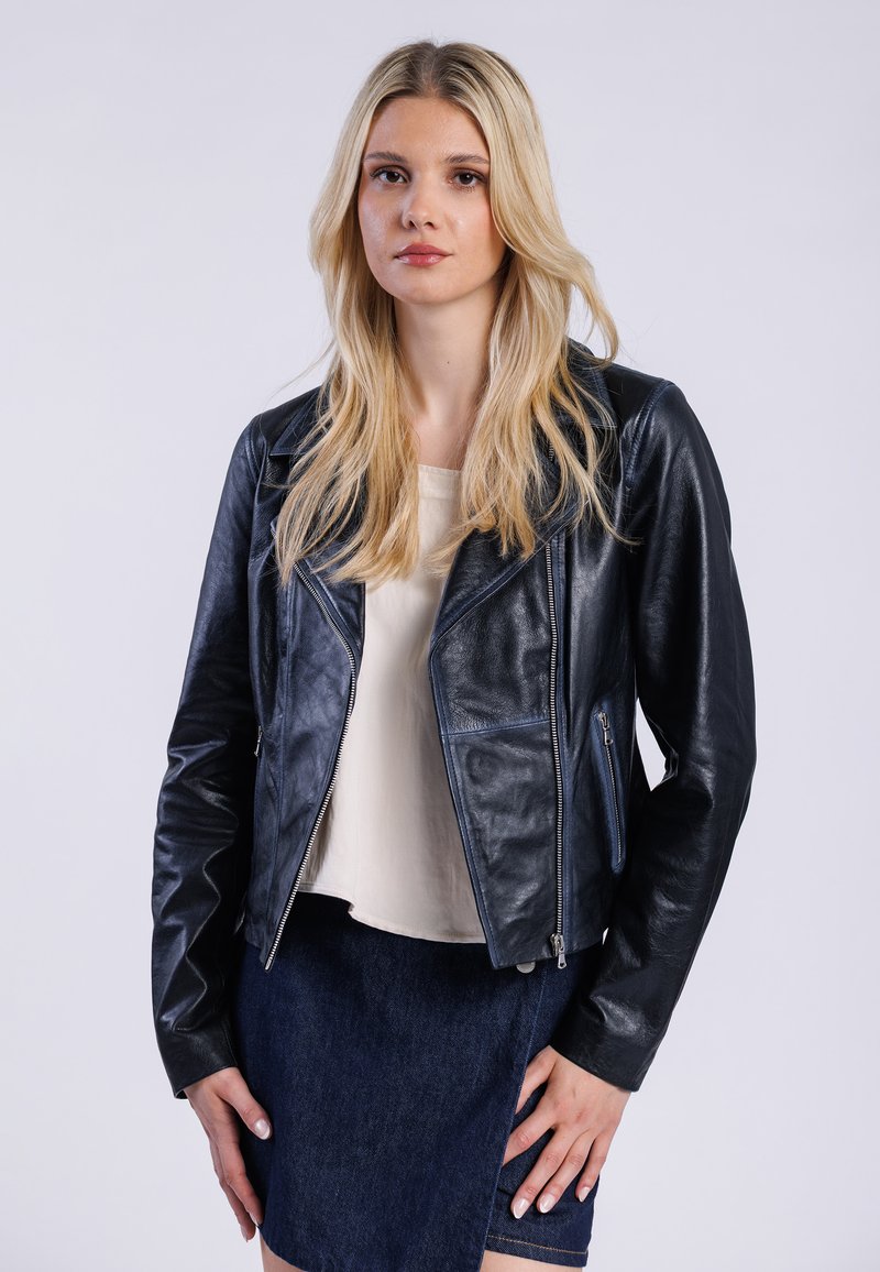 Blondynka w czarnej skórzanej kurtce i jasnej bluzce, stojąca na jednolitym białym tle, z pewnym siebie wyrazem twarzy.