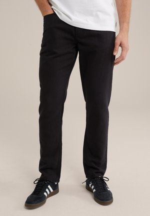 Schwarze Jeans im Slim-Fit, mit einer glatten Textur und dezenten Nähten. Getragen mit schwarzen Schuhen mit weißen Akzenten.