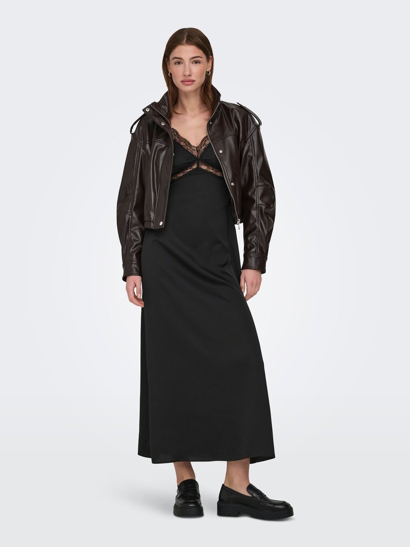 Femme debout portant une robe nuisette noire à dentelle, une veste courte en cuir brun foncé et des mocassins noirs sur un fond blanc uni.